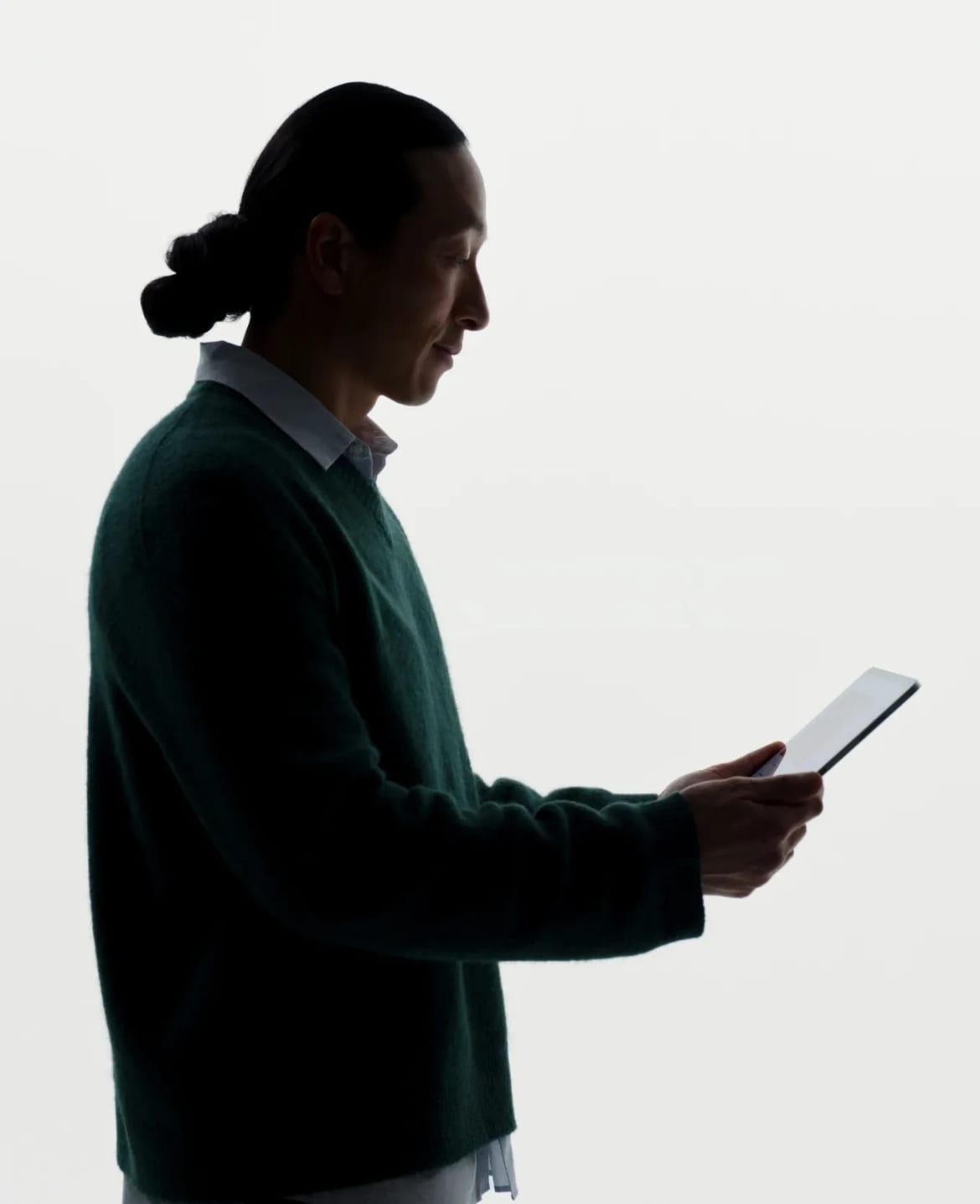 A person in profile in a dark green jumper, reading a tablet, against a white background